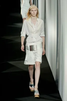 Model in white belted dress walks runway, paired with shiny sandals, during a fashion show.