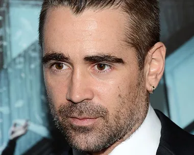 Man with a man-bun hairstyle, wearing a suit and tie, with a serious expression at an event.