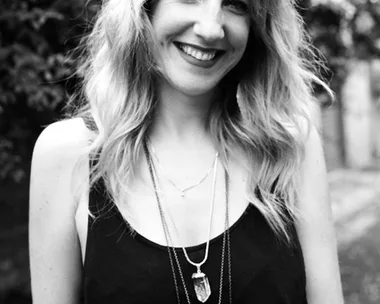 Woman with wavy hair in black tank top, wearing layered necklaces, smiling outdoors; background is blurred foliage.