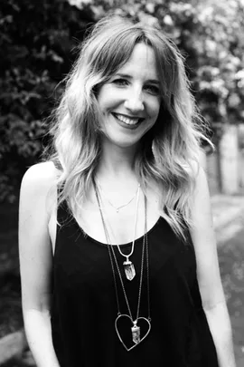 Woman with wavy hair in black tank top, wearing layered necklaces, smiling outdoors; background is blurred foliage.