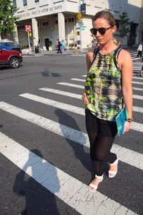 A woman in sunglasses crosses a city street in front of a church, holding a blue clutch and wearing a colorful top.