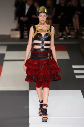 Model wearing an Alexander McQueen SS14 design with a bold leather harness, red skirt, and gold helmet on the runway.