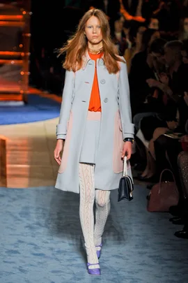 Model in a light blue coat, orange top, white patterned tights, and purple shoes on Miu Miu SS14 runway.