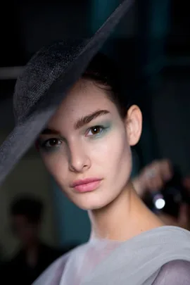 A model wearing a wide-brimmed hat with light blue eye makeup and soft pink lips, posing for a photo.