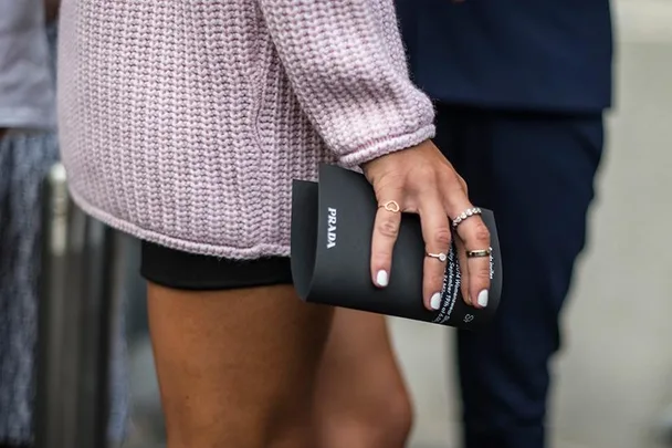 Person in a pink knit sweater holding a black Prada envelope, adorned with rings on their hand.