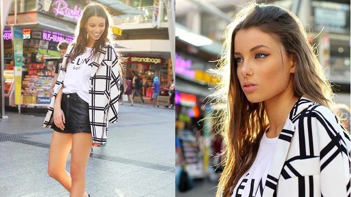 A young woman poses outdoors in a checkered jacket and white t-shirt; the background shows a busy street with colorful signs.