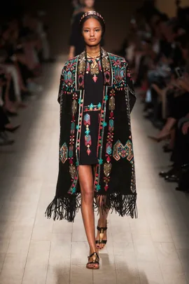 Model walks runway in Valentino SS14 fashion show wearing a black embroidered cape with colorful patterns and fringe.