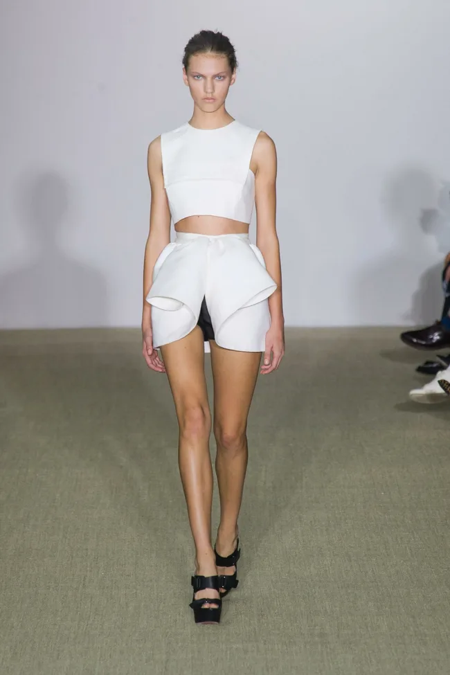 Model wearing a white crop top and structured shorts walks the runway at a fashion show.