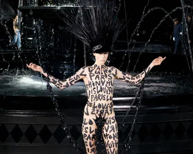 Model in black headpiece and bodysuit with "Louis Vuitton" and "Paris" text, poses near a fountain.