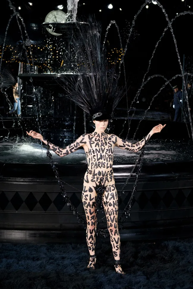 Model in black headpiece and bodysuit with "Louis Vuitton" and "Paris" text, poses near a fountain.