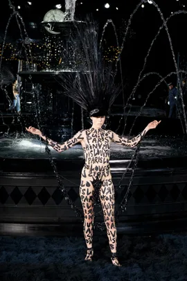 Model in black headpiece and bodysuit with "Louis Vuitton" and "Paris" text, poses near a fountain.