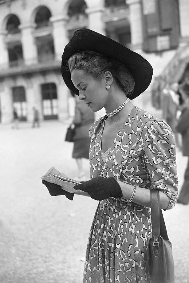 A woman in vintage attire reads a paper, wearing a floral dress, wide-brim hat, pearl necklace, and gloves.