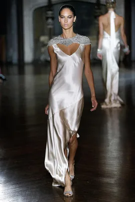 Model walking runway in a silver satin gown with beaded collar, showcasing elegant bridal fashion.