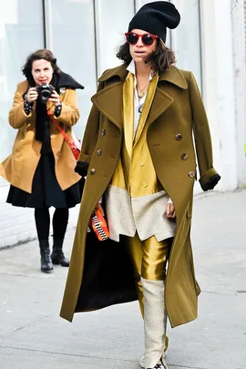 Person in layered fashion: olive coat, mustard suit, black beanie, red sunglasses; photographer in background.