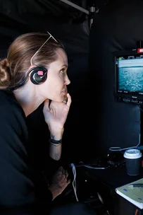 Angelina Jolie on the set of "Unbroken" wearing headphones, observing a screen with an ocean scene.