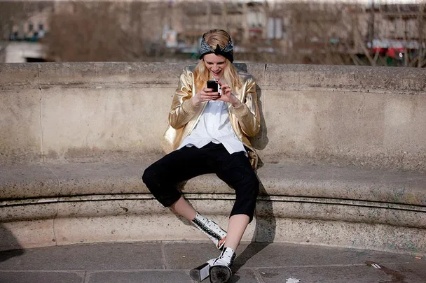 A person in a gold jacket sits on a stone bench, smiling and using a smartphone.