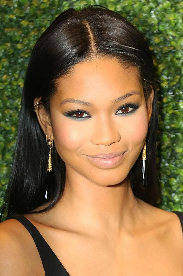 Portrait of Chanel Iman with long black hair and gold earrings, smiling against a leafy background.