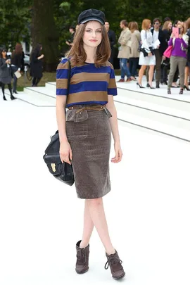 A woman in a striped shirt, skirt, and cap poses outdoors with people in the background at a fashion event.