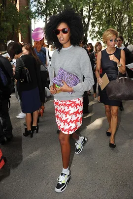 Person in stylish outfit, gray sweater, red patterned skirt, sneakers, and sunglasses, holding a purple bag, walking in a busy street.