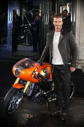 David Beckham in a leather jacket with an orange BMW motorcycle at a nighttime outdoor event.