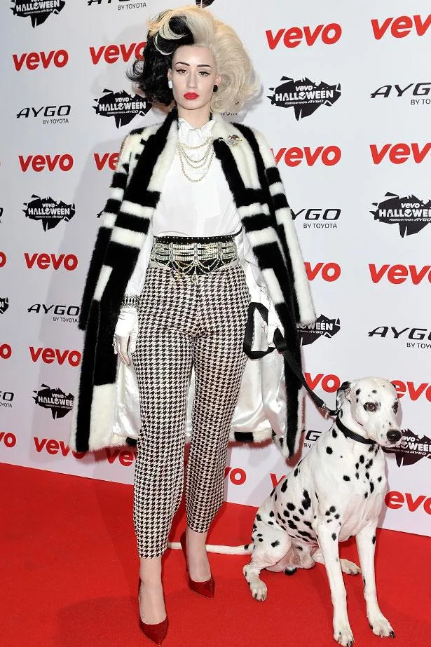 Person dressed as Cruella de Vil with black and white hair, fur coat, and Dalmatian on a red carpet.