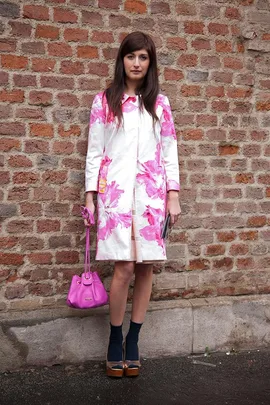 Woman in a white coat with pink floral print, holding a pink bag, standing against a brick wall.