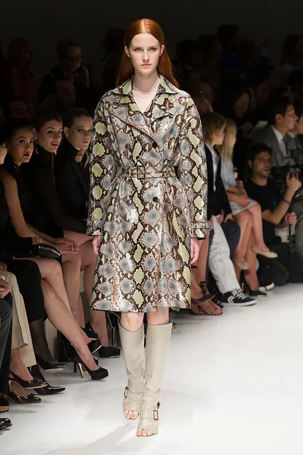 Model on runway in snake-print coat and high boots, audience seated on either side.