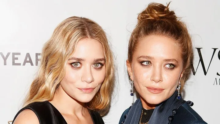 Two women posing together in evening wear at an event, one with blonde hair down and the other with hair in an updo.