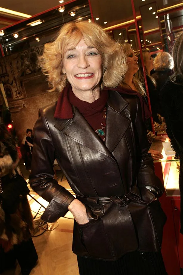 Woman in a brown leather jacket smiling at an indoor event with mirrors and lights in the background.