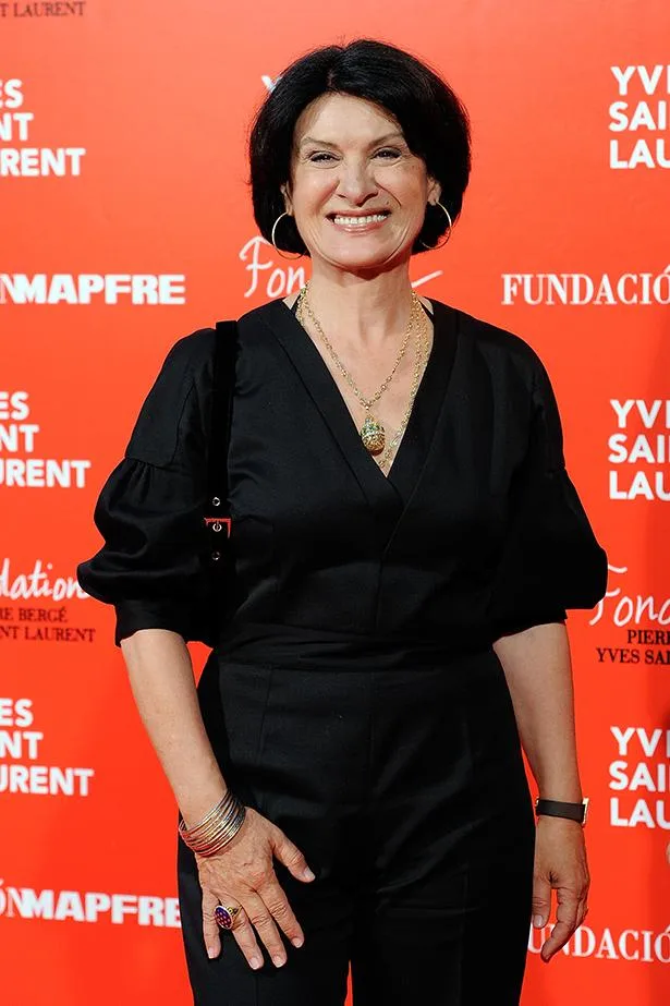 A woman in black outfit smiling at a Yves Saint Laurent event with a red background.
