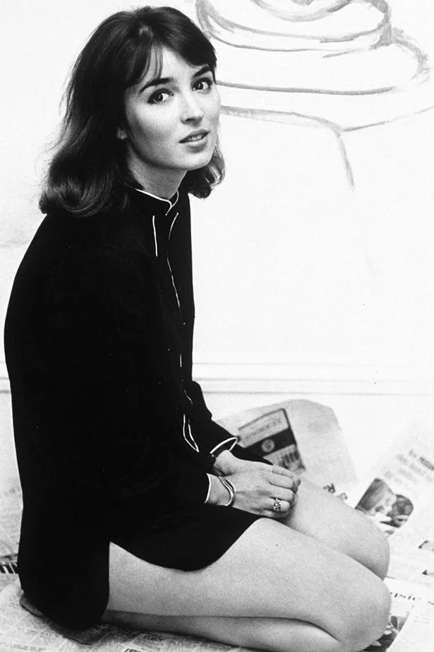 Young woman seated on the floor, looking at the camera, wearing a dark dress with light trim, against a minimalist background.