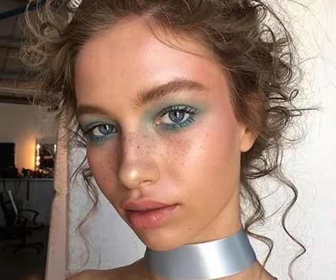 Woman with curly hair wears blue eyeshadow and a silver choker, showcasing a trendy makeup look.