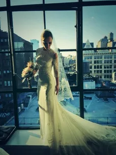 A bride in a lace Givenchy gown and veil stands by a large window overlooking a cityscape, holding a bouquet.