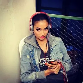 Young woman in denim jacket and headphones holding a phone, sitting against a wall.