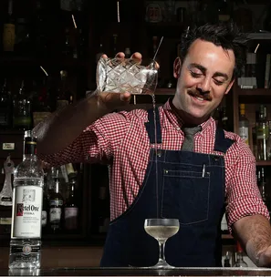 Bartender in checkered shirt and apron pouring a cocktail with Ketel One vodka on a bar counter.