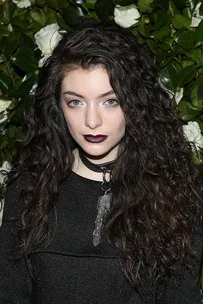 Singer Lorde with long curly hair, wearing dark lipstick and a black outfit, stands in front of a leafy background.