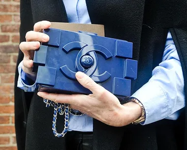 Person holding a blue LEGO-inspired clutch featuring a prominent Chanel logo, wearing a blue shirt and a dark coat.