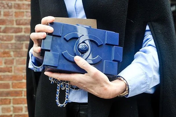 Person holding a blue LEGO-inspired clutch featuring a prominent Chanel logo, wearing a blue shirt and a dark coat.