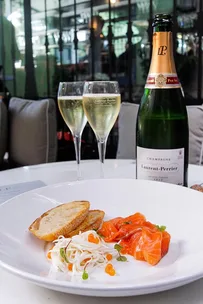 Plate with salmon, bread, and garnish, two filled champagne flutes, and a bottle of Laurent-Perrier Champagne on a table.