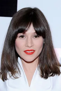 Woman with straight brown hair and blunt bangs, wearing bold red lipstick and a white top, against a light-colored background.