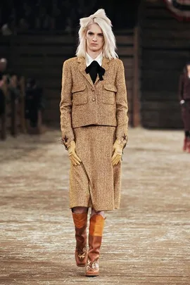 Model in a tan tweed jacket and skirt with a black bow tie, wearing gloves and brown boots on a runway.