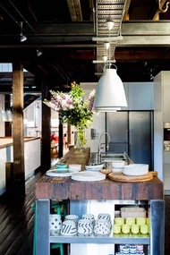 Rustic kitchen with wooden island, open shelves, plates, stylish mugs, and pendant lights; flowers in vase add elegance.