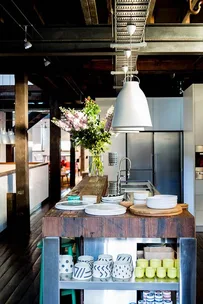 Rustic kitchen with wooden island, open shelves, plates, stylish mugs, and pendant lights; flowers in vase add elegance.