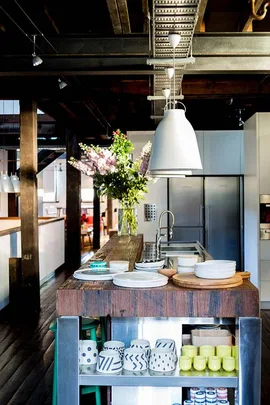 Rustic kitchen with wooden island, open shelves, plates, stylish mugs, and pendant lights; flowers in vase add elegance.