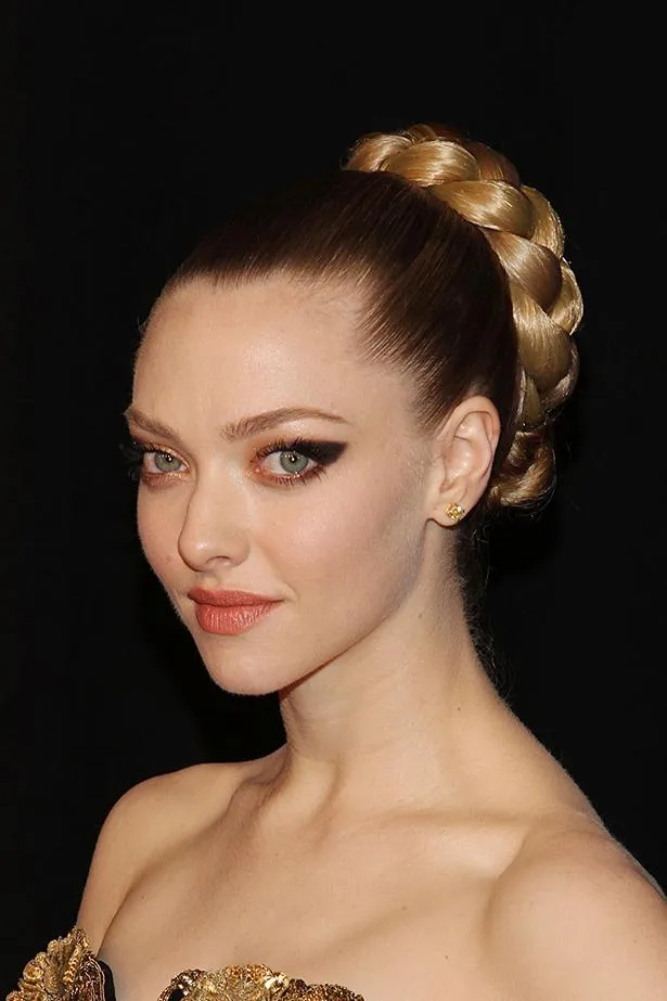 Amanda Seyfried with braided updo and gold dress at a formal event, posing against a black background.