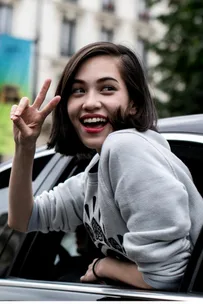 Young woman smiling and making a peace sign from a car window, wearing a gray sweater with printed design.