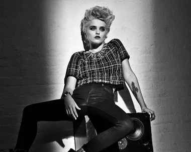 Black and white image of a person with short, styled hair, leaning on a wall with a spotlight, dressed in a checkered top and leather pants.