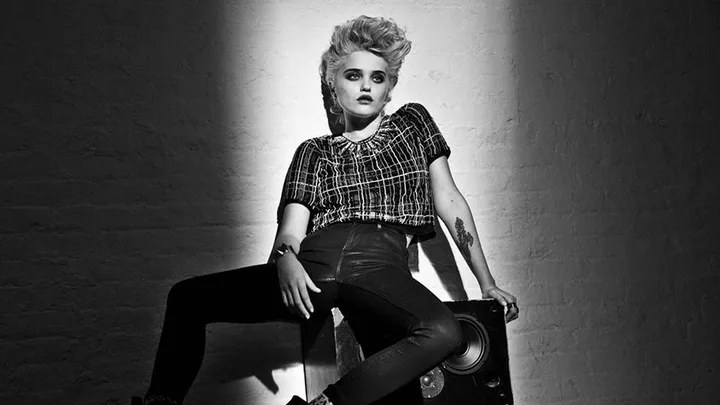 Black and white image of a person with short, styled hair, leaning on a wall with a spotlight, dressed in a checkered top and leather pants.