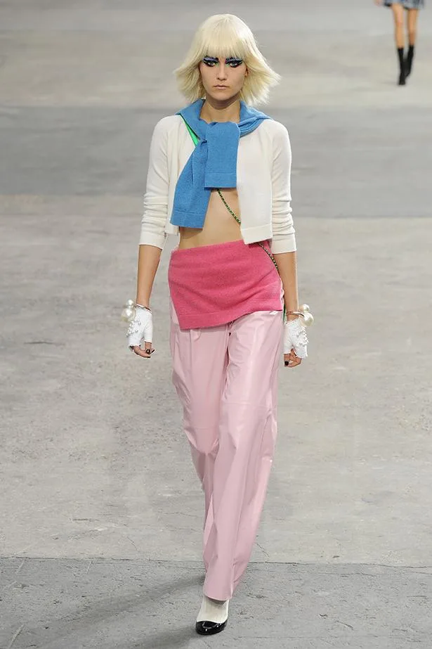 Model walking runway in a white top, pink pants, with blue and pink accessories, white hair, and dramatic eye makeup.