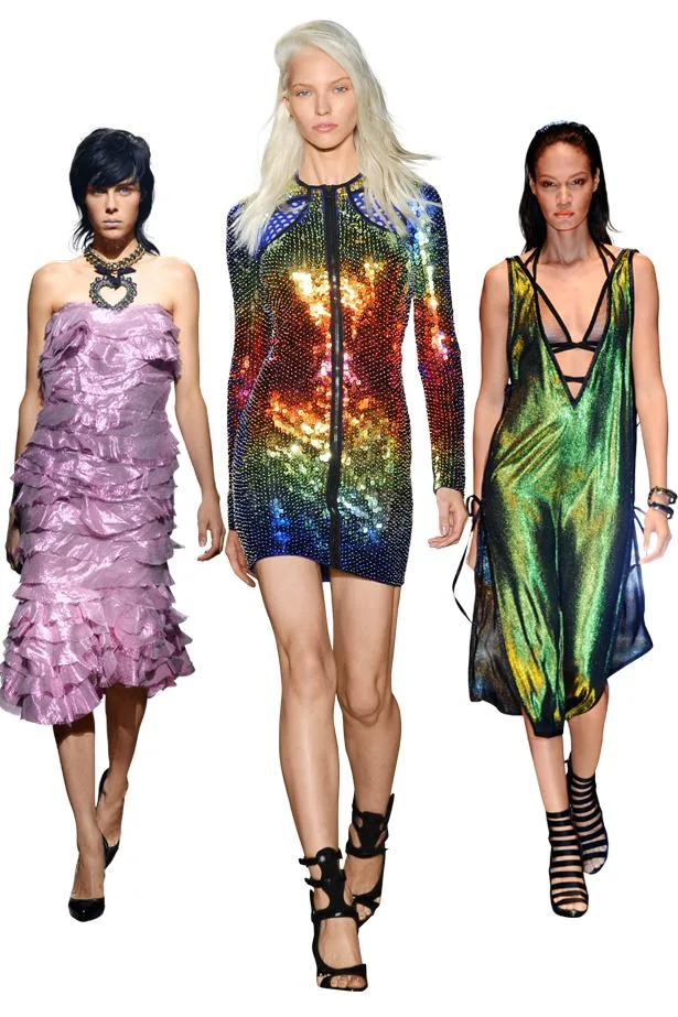 Three models walking runway in colorful, stylish outfits; a pink ruffled dress, a rainbow sequin dress, and a metallic gown.